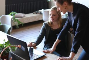 Two professional women discuss a project using a laptop in a modern office environment.