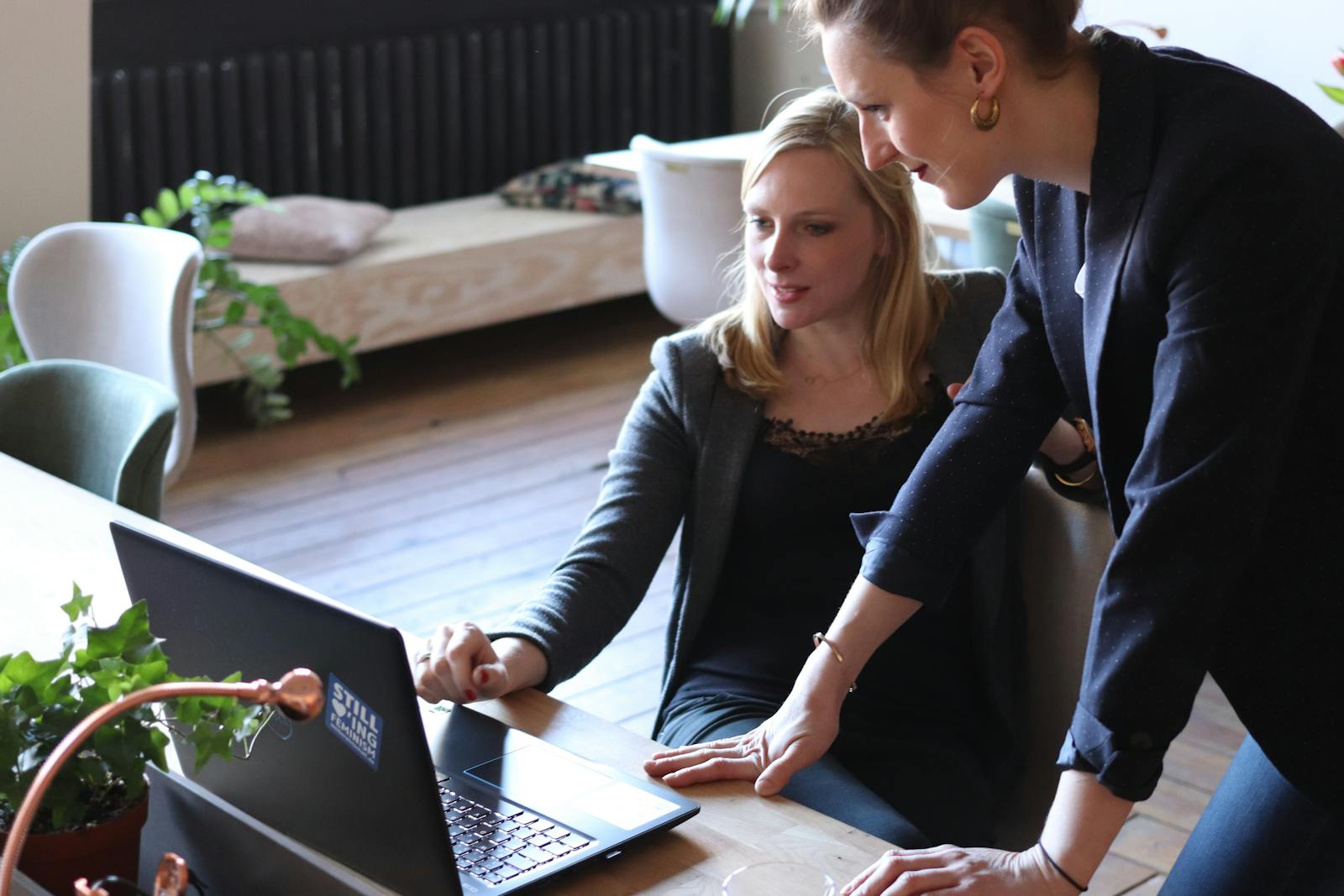 Two professional women discuss a project using a laptop in a modern office environment.