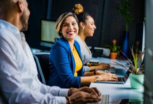 Three diverse professionals working and smiling at office desks, fostering teamwork and collaboration.