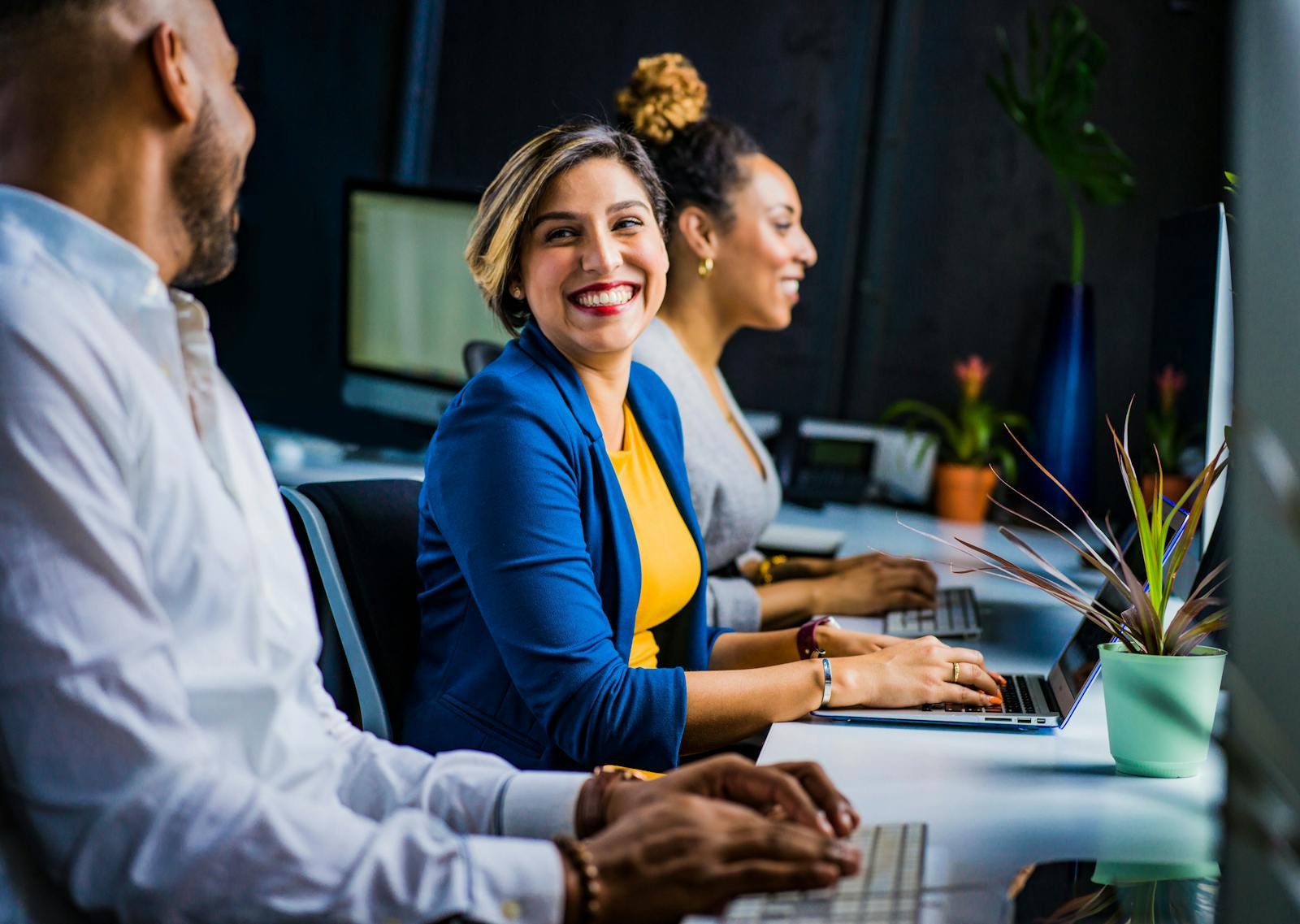 Three diverse professionals working and smiling at office desks, fostering teamwork and collaboration.